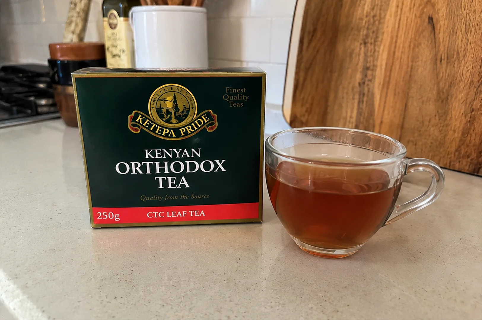 A dark green rectangular box of "Ketepa Pride Kenyan Orthodox Tea" sits on a light-colored, speckled kitchen countertop next to a clear glass mug filled with amber-colored brewed tea. The tea box features a gold and black circular logo with the brand name on a gold ribbon, bold white lettering reading "KENYAN ORTHODOX TEA," and a red stripe across the bottom indicating "250g CTC LEAF TEA." In the background, the soft-focused kitchen setting shows a wooden cutting board leaning against a white tiled backsplash, along with a white ceramic utensil holder and a bottle of olive oil.
