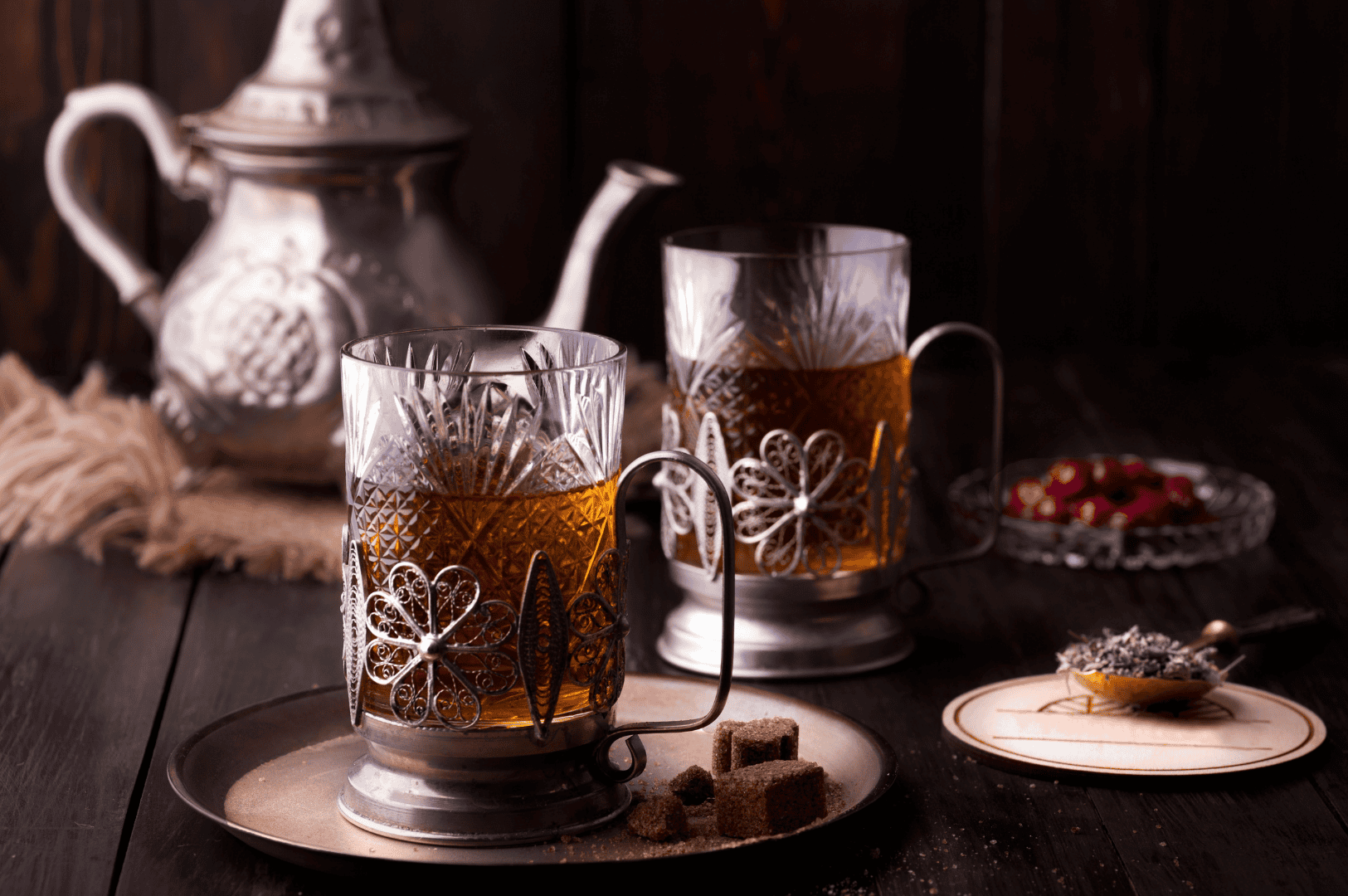 Elegant Ceylon high grown tea service inspired by Nuwara Eliya, featuring two ornate glass holders with amber tea on a vintage metal tray, brown sugar cubes, silver teapot, red berries, and dried lavender on a dark wooden table with warm, moody lighting.
