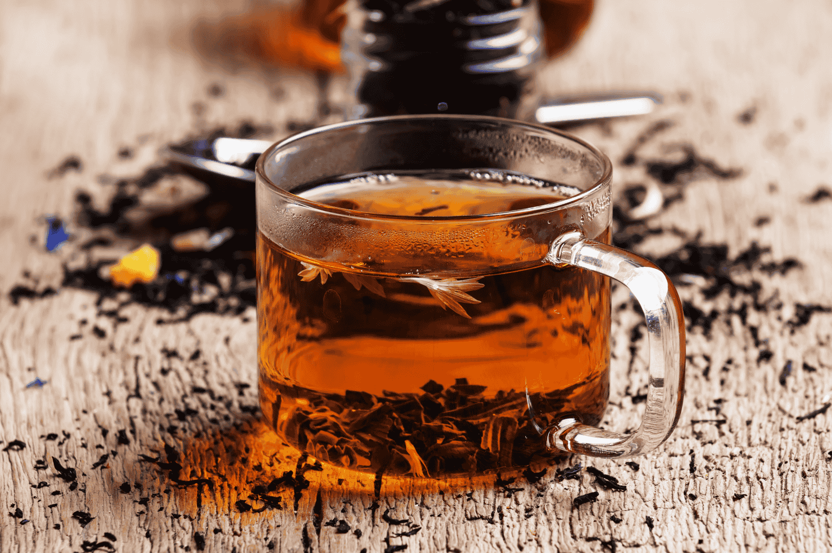Clear glass cup of amber Ceylon high grown tea from Nuwara Eliya, infused with loose leaves and white blossoms, surrounded by scattered tea leaves and petals on a rustic wooden surface, evoking the region’s bright, crisp flavor profile.