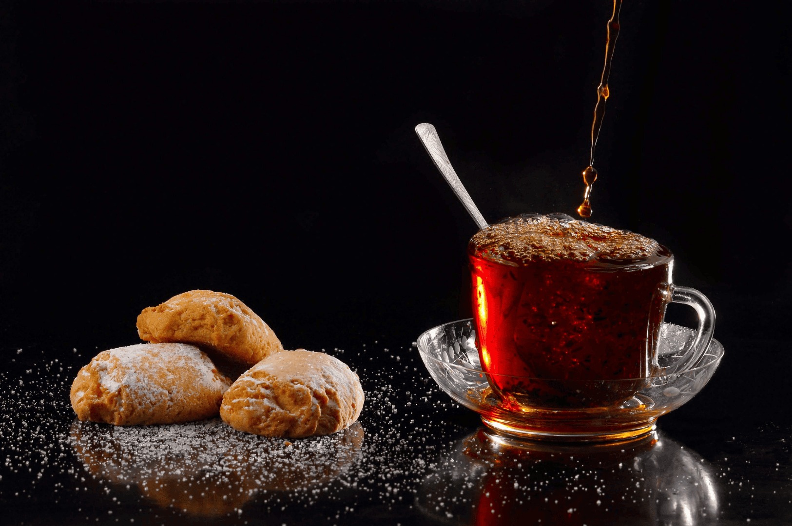 Amber Ceylon high grown tea from Nuwara Eliya being poured into a clear glass cup with a metal spoon, served alongside powdered sugar‑coated cookies on a dark reflective surface, highlighting the region’s bright, crisp flavor profile.