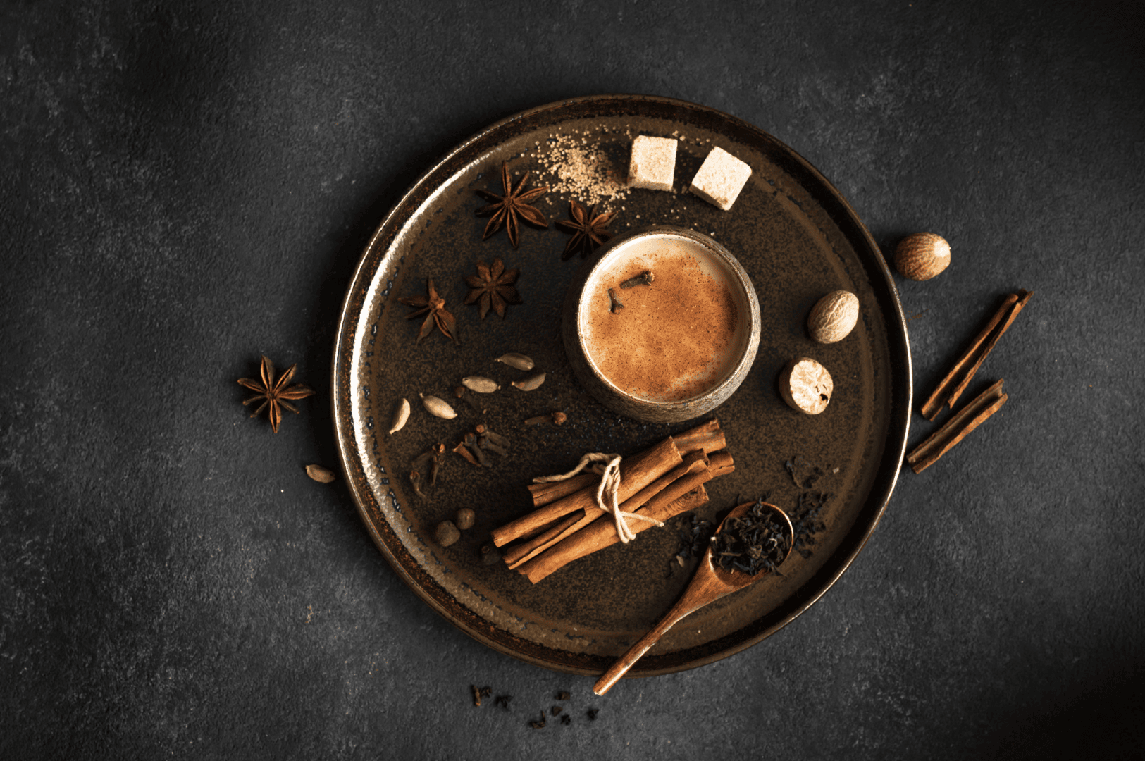 Rustic plate with a cup of masala chai (chai tea) topped with froth, surrounded by whole spices including cinnamon sticks, star anise, green cardamom pods, nutmeg, cloves, and a wooden spoon of tea leaves. This spiced tea blends black tea, darjeeling tea, assam tea, or green tea with fresh ginger, ground ginger, chai masala powder, tea masala, and fragrant spices from tea plantations in South India. Rooted in indian masala chai traditions, masala chai translates to traditional Indian tea brewed with milk and sugar, oat milk, or steamed milk, sweetened with brown sugar, coconut sugar, or maple syrup. Whether enjoyed as a chai tea latte, chai latte, milky tea, or milky cup, it’s a hot cup of comfort for tea drinkers and indian tea drinking culture. Made with loose tea, black tea bags, or ctc tea, this masala tea is tea brewed with aromatic spices, spice mix, and ground spices using a spice grinder. From tea stalls, local coffee shop, and coffee shops to south asian grocery stores, tea vendors, tea sellers, and chai vendors, the tea trade, tea board, and tea production keep tea produced traditions alive. This perfect cup celebrates indian tea culture, make masala chai heritage, and why this recipe endures across the Indian subcontinent, from tea plant origins to drinking tea moments and tea breaks enjoyed worldwide, infused with aromatic herbs, fennel seeds, ceylon cinnamon, and all the spices.