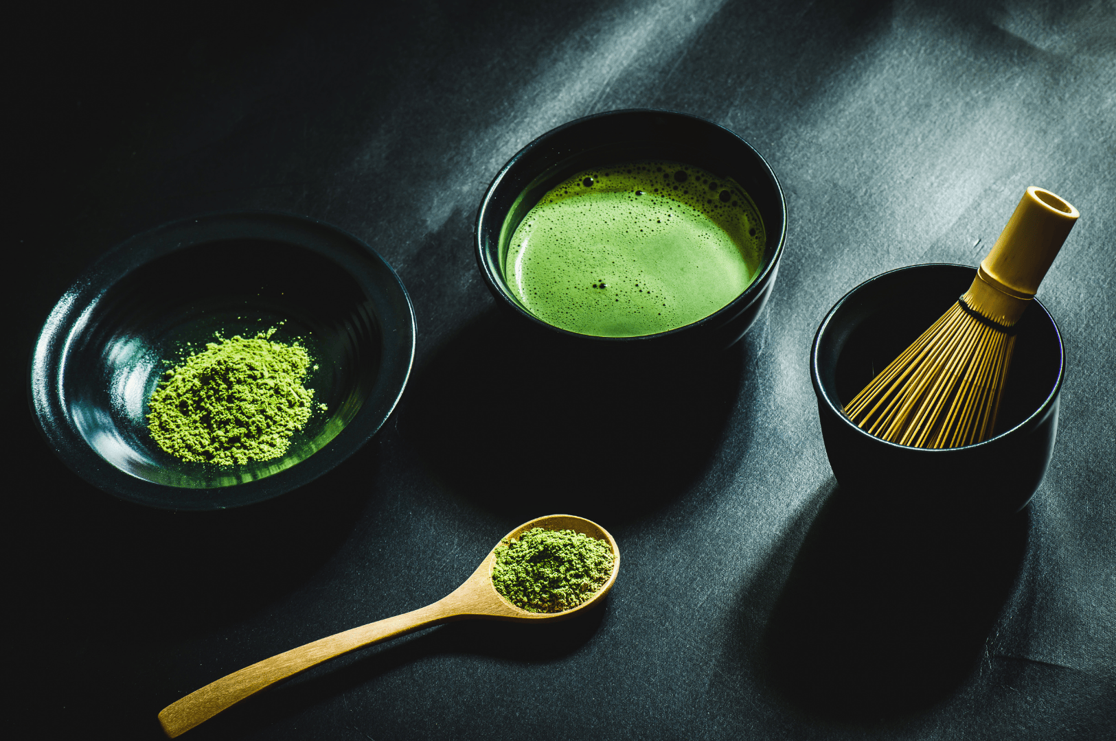 Traditional matcha tea preparation setup featuring ceremonial grade matcha powder in a black dish, a bamboo whisk (chasen), and a bowl of frothy ceremonial matcha made with hot water, showcasing the vibrant green color and fine texture of high quality matcha. This scene reflects tea ceremonies in Japan, where tea masters use stone ground matcha powder from tea leaves and tea plants harvested during the first harvest and shade-grown under sunlight. Matcha grades—from culinary grade matcha to superior ceremonial matcha—are determined based on particle size, form, flavor, and quality. Naoki matcha, a popular japanese brand, offers different grades including highest grade ceremonial grade and culinary grade options for drinking, baking, cooking, desserts, smoothies, and matcha lattes with milk, sugar, or sweetener. Good matcha delivers umami taste, strong flavor, and a balance of bitterness and sweet notes, unlike coffee. Not all matcha is equal—green tea varieties like sencha and japanese green tea differ in taste, powder quality, and how they’re mixed with other ingredients. This image celebrates the artistry of japanese tea culture and the ritual of preparing higher quality beverages with ground matcha.