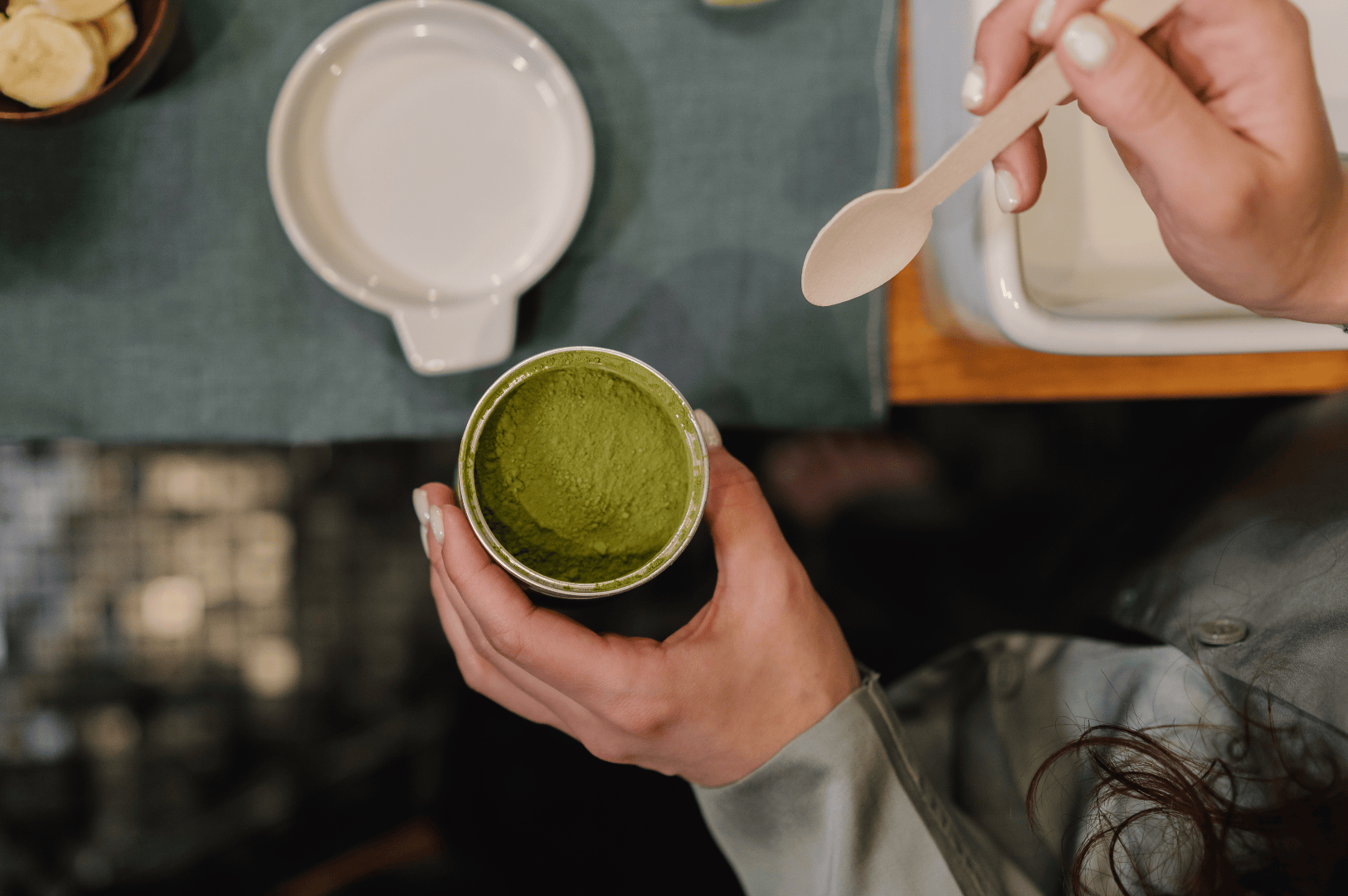 Person scooping vibrant green matcha powder from a container using a wooden spoon, preparing ceremonial grade matcha for matcha lattes and desserts. Surrounded by sliced bananas, a ceramic bowl, and a green tablecloth, this scene highlights the vibrant green color and umami taste of superior ceremonial matcha and culinary grade matcha. Featuring naoki matcha, a high quality japanese green tea brand, the image evokes traditional tea ceremonies led by tea masters using stone ground grade matcha powder from first harvest tea leaves. Not all matcha is equal—different grades like culinary grade and ceremonial grade are determined based on particle size, shade, and flavor. Matcha grades reflect the quality of japanese tea produced from steamed tea plants grown in sunlight-limited conditions in Japan. Good matcha offers strong flavor, smooth texture, and a sweet, slightly bitter taste ideal for drinking, baking, cooking, smoothies, and beverages. Mixed with hot water, milk, sugar, or sweetener, matcha rivals coffee in versatility. Whether used in sencha-style green tea, desserts, or lattes, this highest grade matcha form showcases the essence of green tea culture and the refined flavor of high quality matcha.