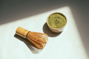 A bamboo whisk (chasen) lies beside a bowl of vibrant green matcha powder, illuminated by sunlight—symbolizing the essence of japanese tea ceremonies and the artistry of tea masters. This image showcases ceremonial grade matcha and culinary grade matcha, highlighting the difference between superior ceremonial matcha and culinary grade matcha powder used in matcha lattes, smoothies, desserts, and baking. Naoki matcha and other high quality matcha brands are produced from shade-grown tea plants and first harvest tea leaves in Japan, offering an umami taste, strong flavor, and smooth texture. Stone ground powder with fine particle size determines matcha grades, from highest grade ceremonial matcha to culinary grade used in cooking and mixed beverages. Not all matcha is equal—good matcha has a vibrant green color, low bitterness, and is often steamed and sweetened with sugar or milk. Whether drinking japanese green tea or green tea lattes, the flavor, form, and quality are determined based on grade matcha powder. Matcha, sencha, and other green teas differ in taste, bitterness, and use in food, coffee alternatives, and sweet or savory recipes. This image evokes the ritual of hot water preparation, the role of the whisk, and the cultural significance of japanese tea, highlighting how matcha is ground, mixed, and enjoyed across different grades and applications.