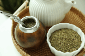 Traditional yerba mate setup featuring a wooden gourd with metal rim and bombilla, filled with dried mate leaves, alongside a ceramic bowl of loose yerba mate and a white teapot on a woven tray—evoking Argentina’s timeless ritual and the modern revival of its energizing brew.