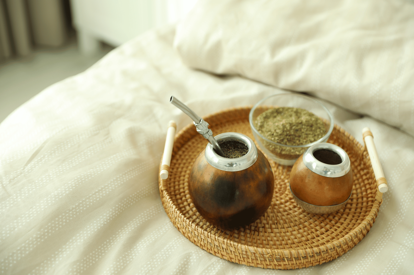 Cozy tray on a bed with light sheets, holding two polished mate gourds—one with a bombilla—and a glass bowl of loose yerba mate leaves, evoking a relaxed, modern take on Argentina’s traditional tea ritual.