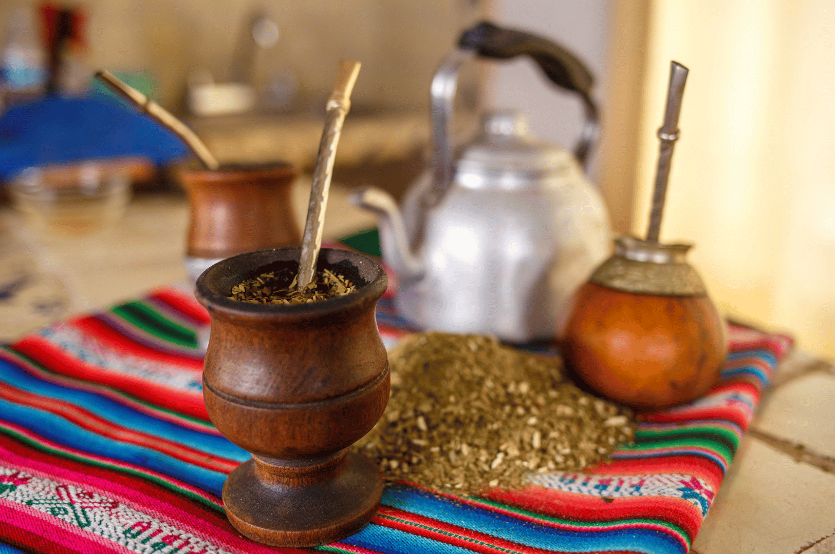 Three traditional mate gourds—wooden and calabash—each with a bombilla, rest on a vibrant Andean textile alongside a metal kettle and loose yerba mate leaves, capturing the communal spirit and ancestral roots of Argentina’s energizing tea ritual.