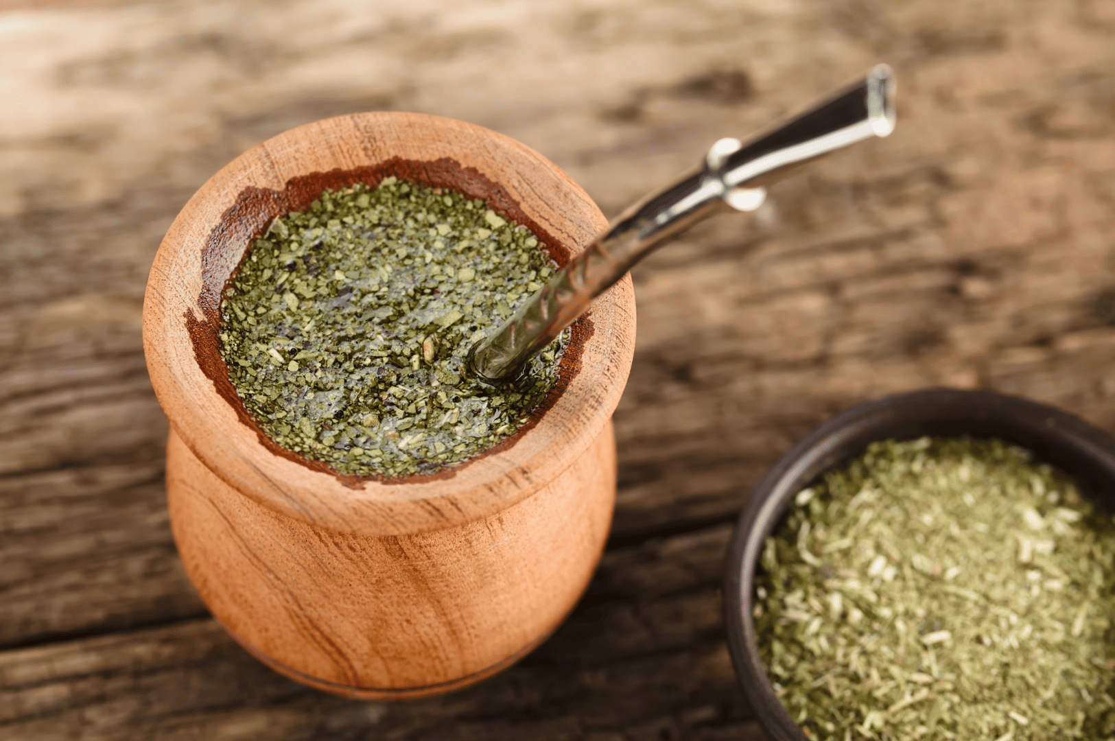 Rustic wooden mate gourd with bombilla, filled with dried yerba mate leaves, beside a small bowl of loose tea on a weathered wooden surface—evoking Argentina’s ancestral energy ritual and the quiet power of tradition.