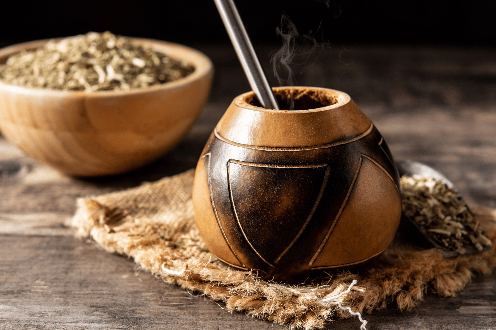 Steaming yerba mate in a carved gourd with bombilla, set on burlap atop a wooden surface, with loose leaves and a metal spoon nearby—evoking the warmth, texture, and meditative ritual of Argentina’s ancient energy brew.