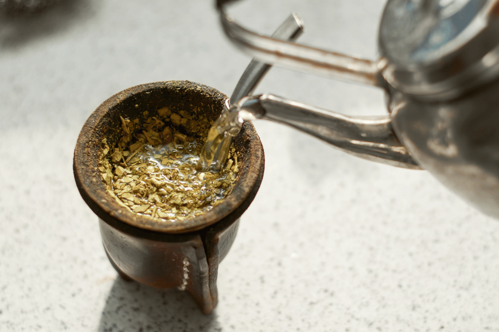 Hot water pouring from a metal kettle into a yerba mate-filled gourd with bombilla, as steam rises and leaves begin to infuse—illustrating the ceremonial start of Argentina’s energizing tea tradition.