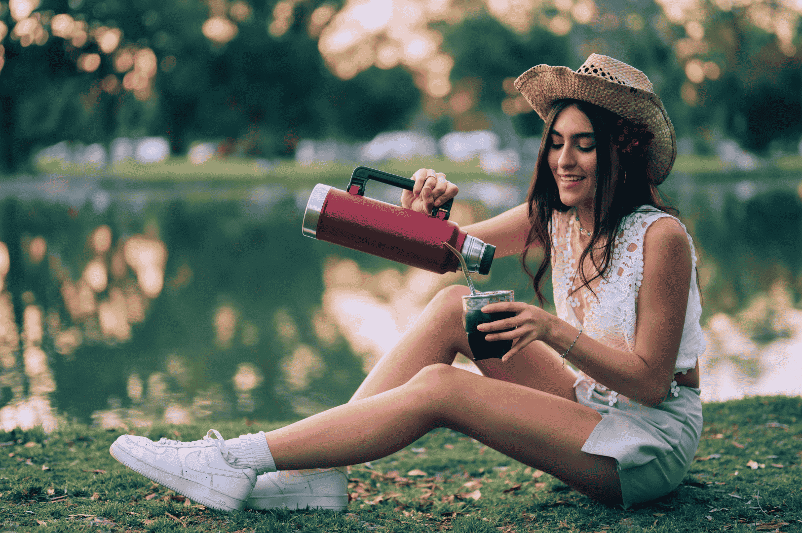 Smiling person in a straw hat pours yerba mate from a red thermos into a glass cup while sitting on grass near a lake—capturing a serene moment of solo enjoyment and the modern, nature-infused revival of Argentina’s traditional energy brew.