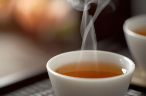 A close-up shot of a small, white Gongfu tea cup filled with amber-colored tea, with wisps of steam rising from the hot beverage.