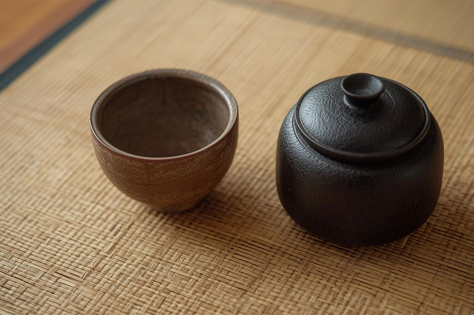 A small, rustic brown ceramic tea bowl (chawan) next to a black, lidded tea caddy (natsume) sitting directly on a woven tatami mat.