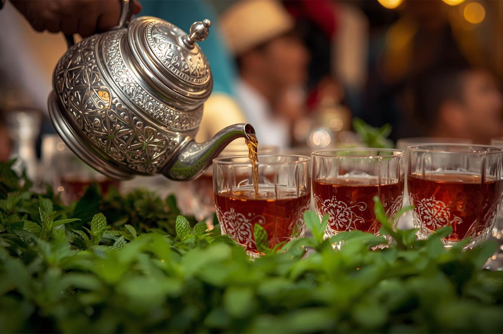 A traditional ornate silver Moroccan teapot pouring tea into small, decorated glass cups, framed by a layer of fresh mint leaves in the foreground.