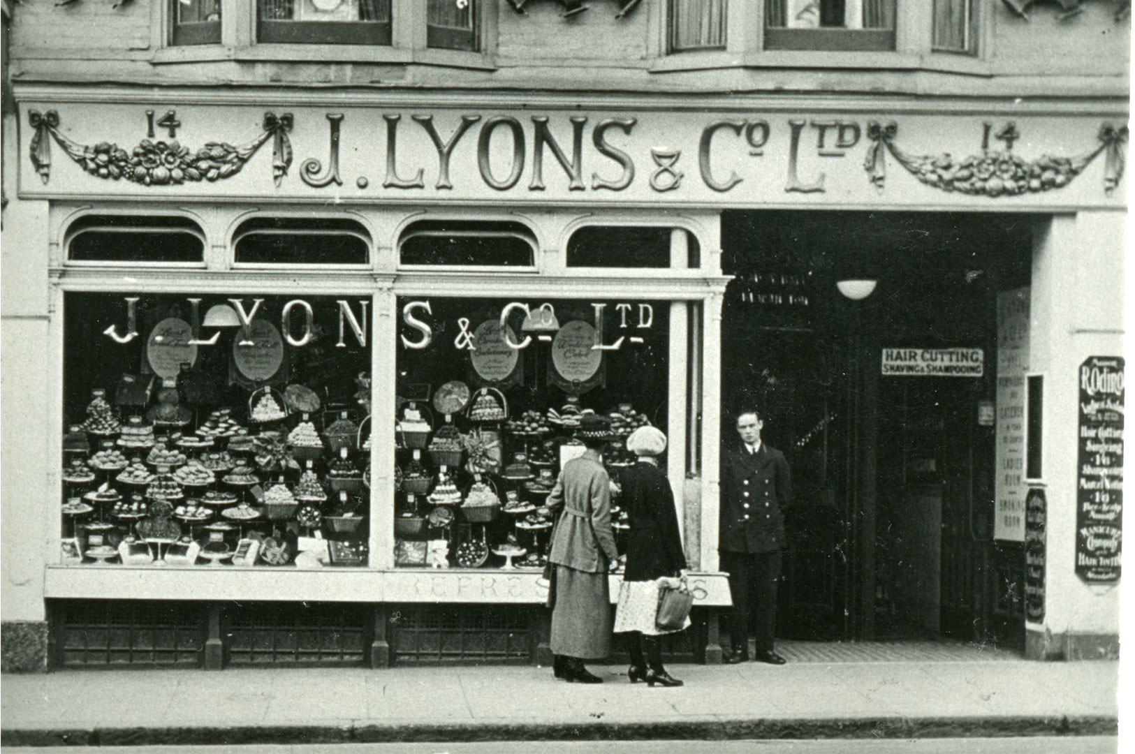A black and white photograph of the storefront of a vintage J. Lyons & Co Ltd tea shop, showing two women looking into the window display of cakes and pastries, and a man in uniform standing by the door.
