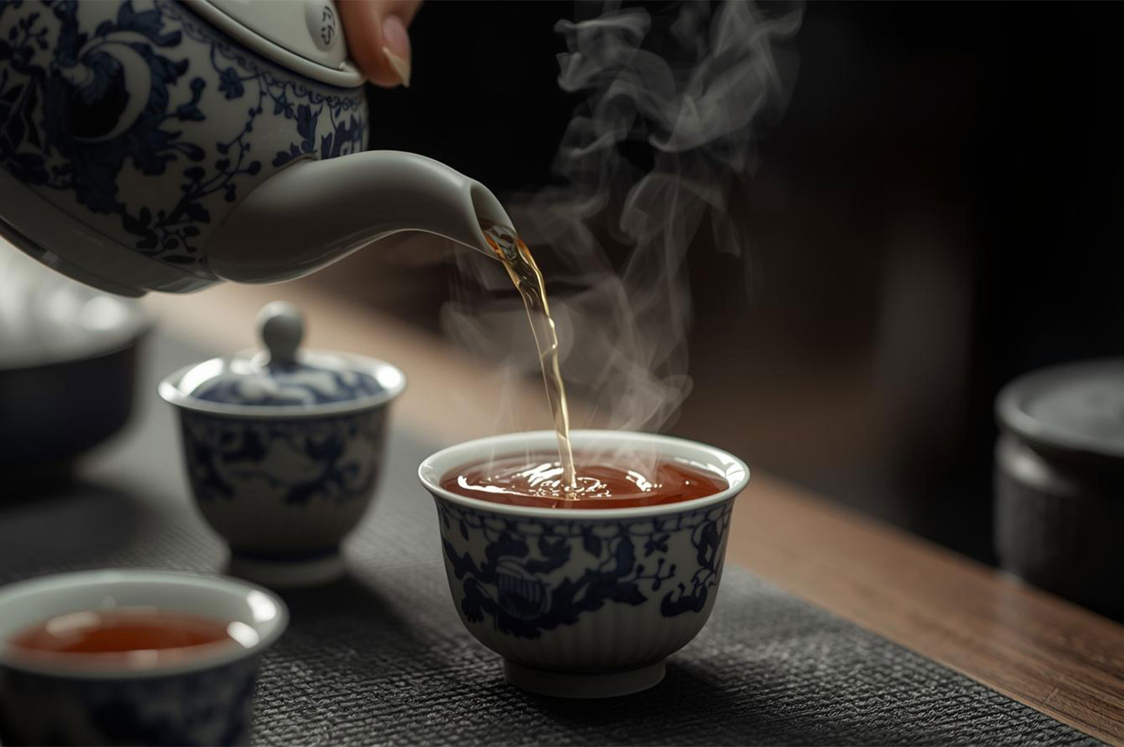 A hand pours hot, dark-amber tea from a blue and white patterned porcelain teapot into a matching teacup, with dramatic steam rising. The scene suggests a traditional Chinese tea ceremony.