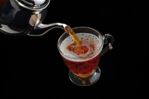 A high-contrast studio shot of hot, amber-colored tea being poured from a silver teapot into a clear glass cup on a black background.