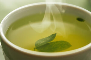 Close-up of a steaming hot cup of light green tea, with two floating leaves and visible steam rising from the surface.