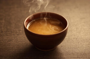 Close-up of a steaming, milky, brownish-yellow beverage, likely Tibetan butter tea (Po Cha), served in a small brown ceramic bowl on a dark wooden table.
