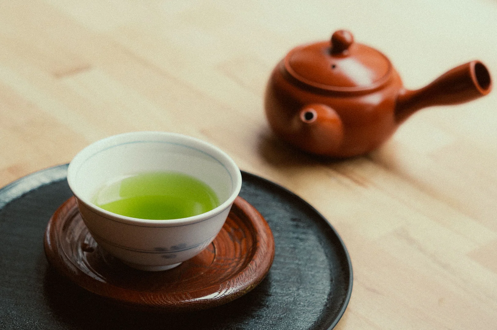 2 - The Complete Guide to Drinking Green Tea A high-angle, clean photograph shows a white ceramic teacup filled with vibrant, bright green matcha tea sitting on a dark wooden saucer, which rests on a larger black circular tray. In the softly blurred background, a traditional reddish-brown clay teapot (kyusu) with a side handle sits on a light-colored wooden table. The focus remains sharp on the teacup in the foreground, highlighting the rich color of the green tea and the subtle blue floral pattern painted on the outside of the white ceramic cup.