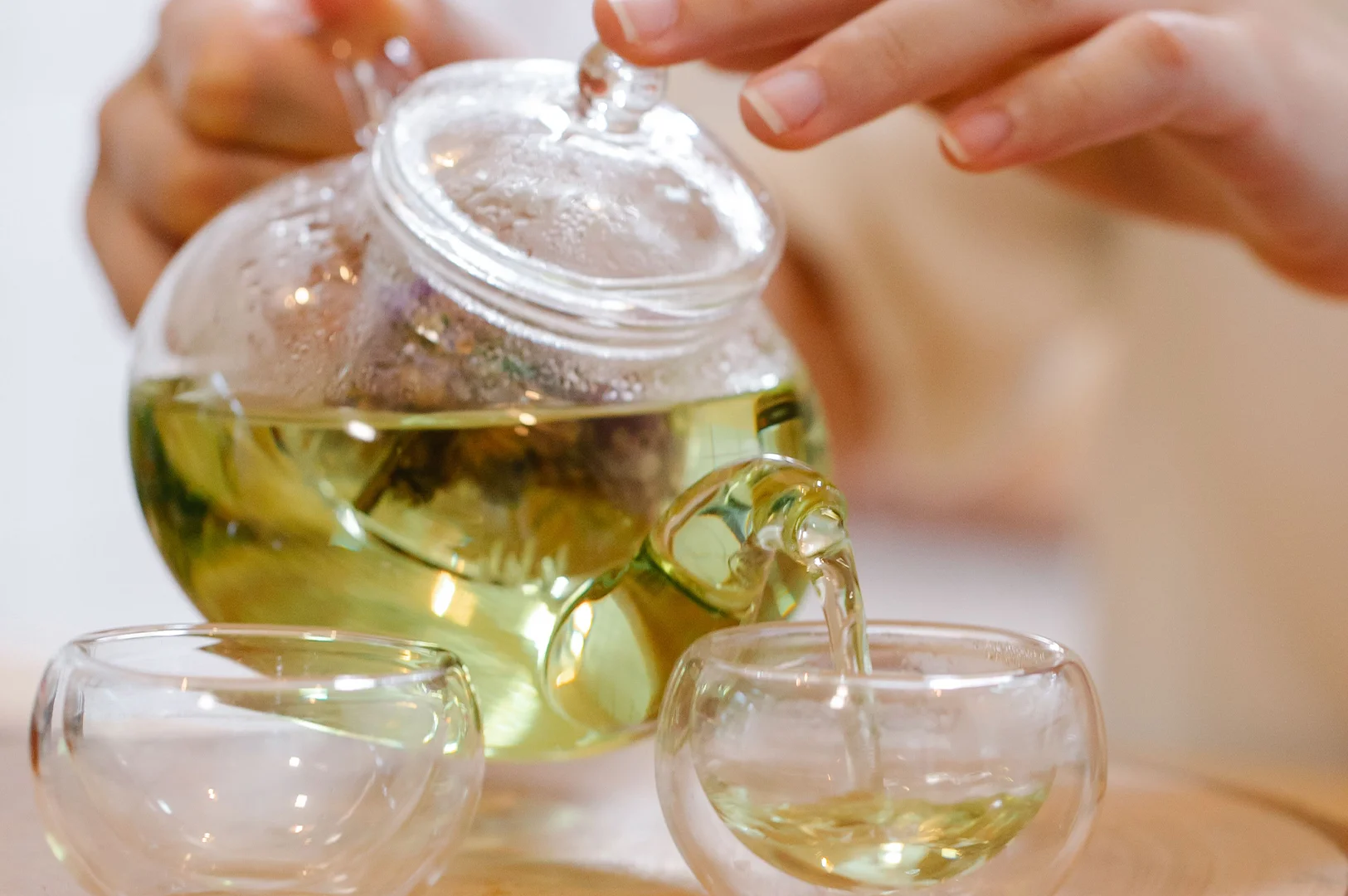 6 - The Complete Guide to Drinking Green Tea A close-up, slightly warm-toned photograph captures a person's hands pouring light-green herbal tea from a clear glass teapot into one of two small, double-walled glass cups resting on a light wooden surface. The person's hands are softly blurred in the background, with one hand gently holding the teapot's round lid while tilting it to pour. Inside the transparent teapot, dark tea leaves or flowers are visible in a mesh infuser, steeping in the golden-green liquid. A steady stream of tea flows smoothly from the curved glass spout into the right-hand cup, which is already partially filled, while the identical cup on the left stands empty and waiting.