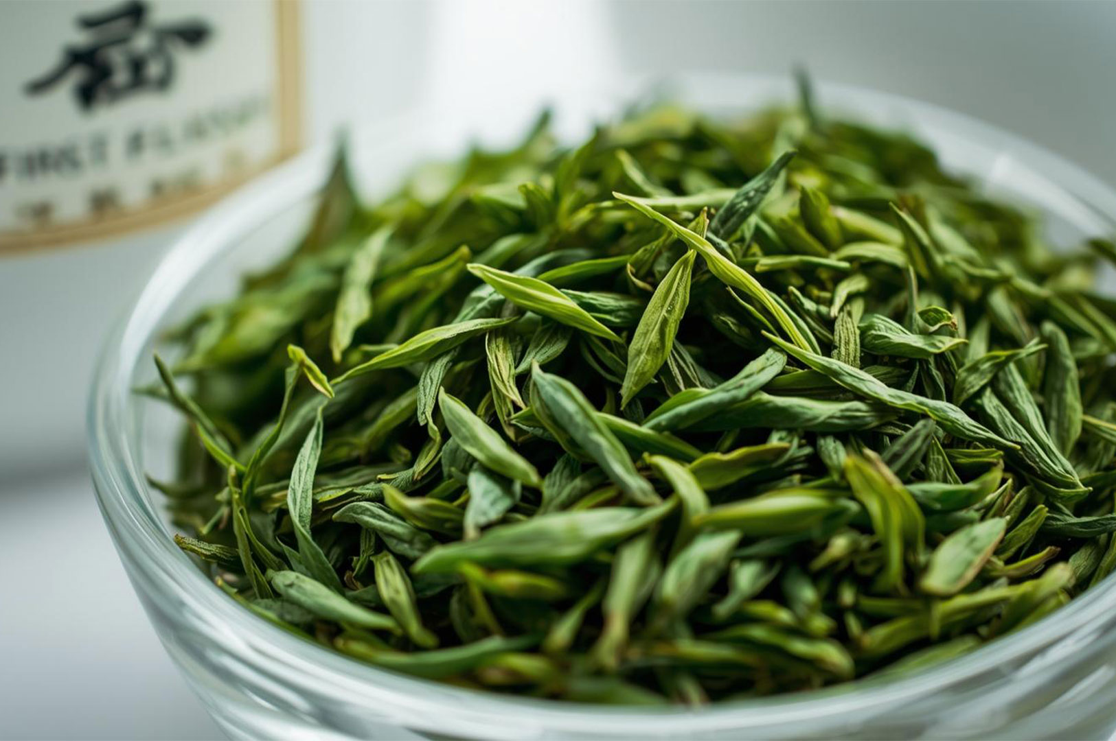Macro close-up of a glass bowl filled with vibrant, high-quality, whole-leaf green tea (likely Longjing or Dragon Well), showing the fresh, spear-like shape of the leaves.