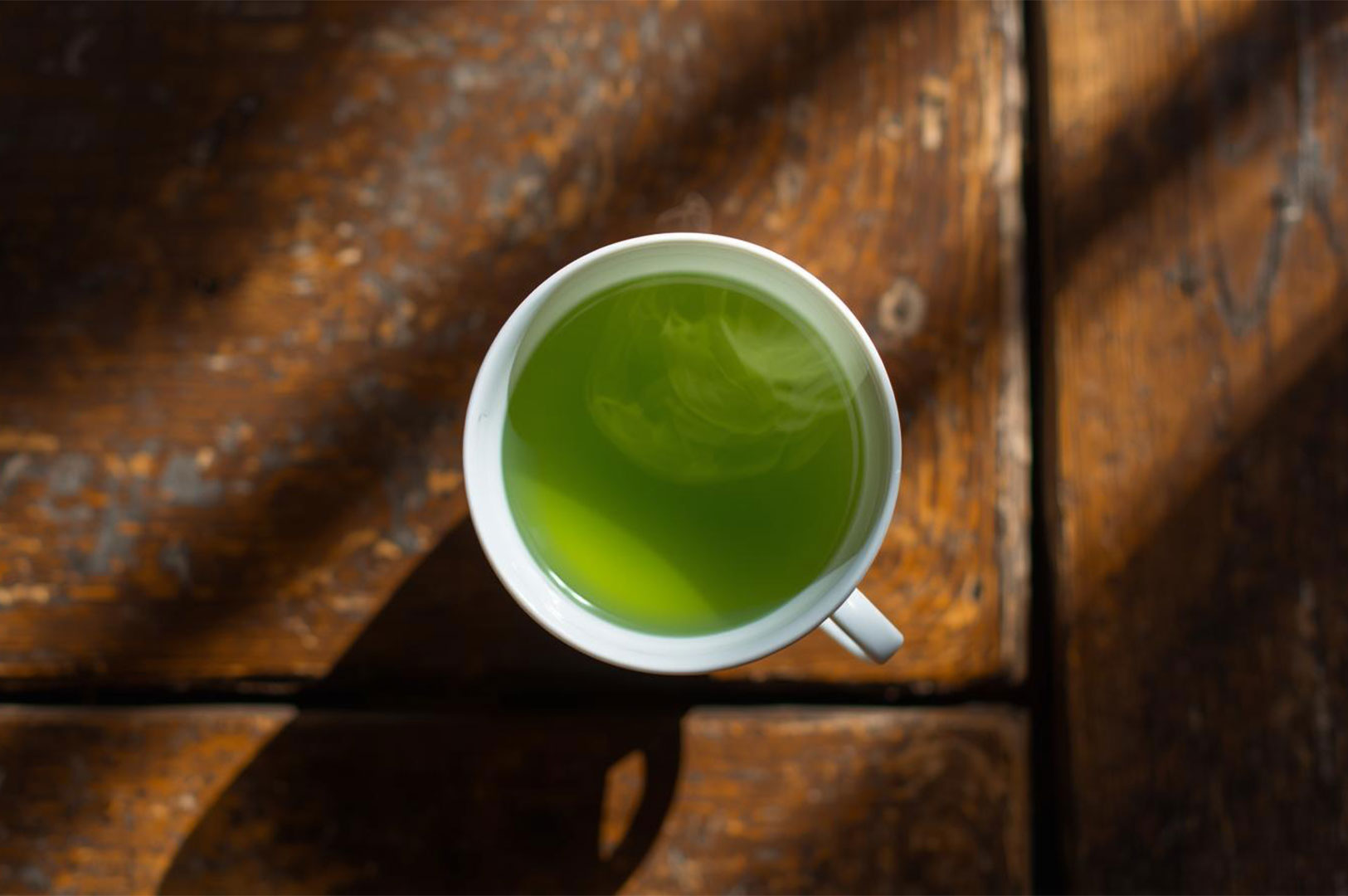 Overhead shot of a white ceramic cup filled with bright green tea (likely matcha) sitting on a rustic wooden table, with dark shadows and steam rising softly from the surface.
