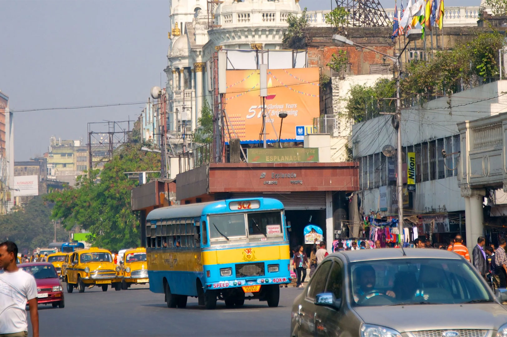 A busy street scene in Kolkata, India, featuring traffic including a prominent blue and yellow public bus, classic yellow ambassador taxis, and various other vehicles, set against a backdrop of historic buildings and street vendors.