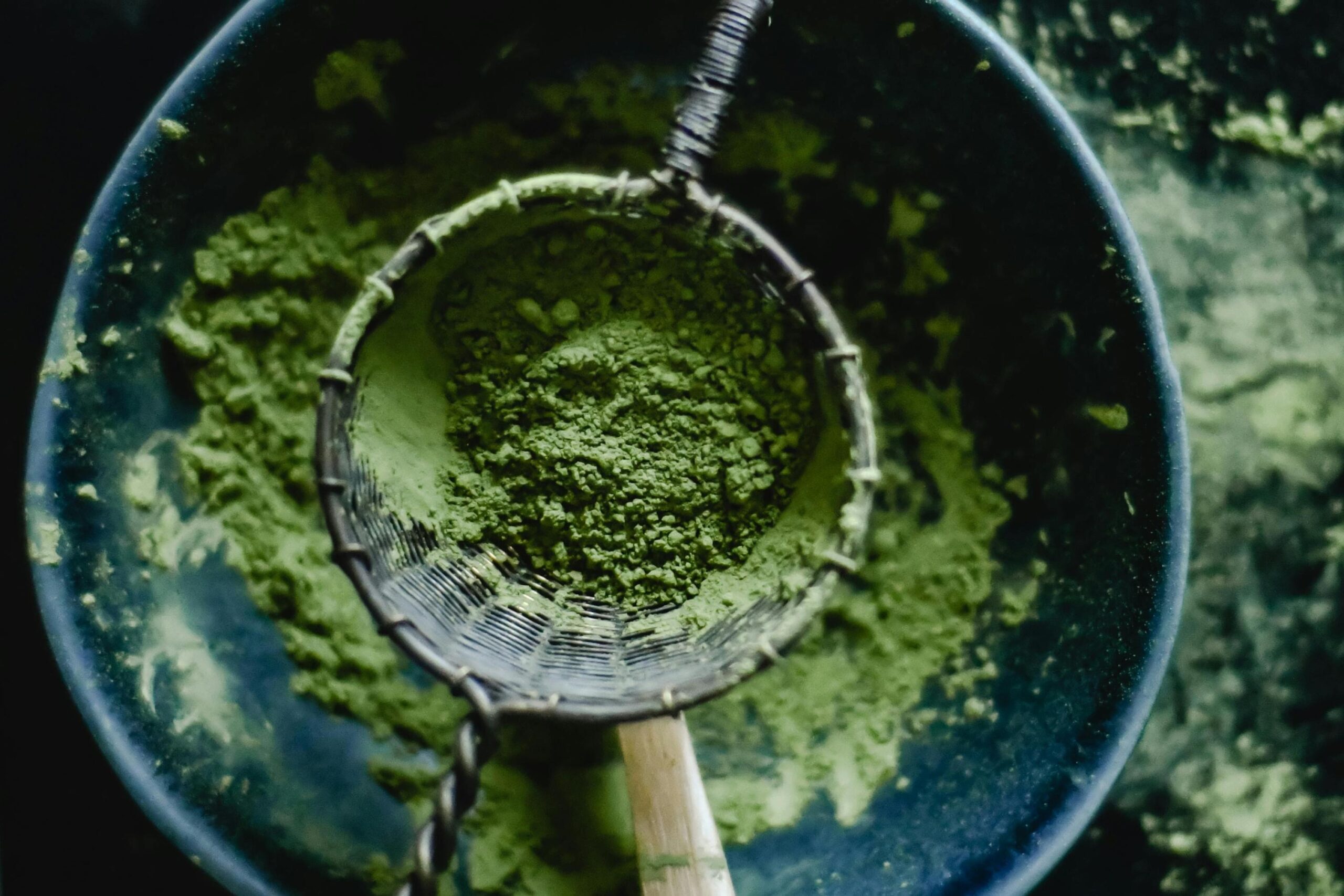 This top-down shot features a dark ceramic bowl filled with fine, vibrant green matcha powder. A metal mesh sifter rests over the bowl, containing a small pile of the concentrated tea powder ready to be filtered.