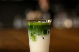 A close-up, eye-level shot of an iced matcha latte served in a clear glass, sitting on a dark wooden table. The drink features a striking layered effect, with a thick layer of creamy white milk and ice cubes at the bottom, topped by a dense, vibrant green matcha layer that gently swirls down into the milk. The top of the beverage has a thin layer of fine green foam. The background is softly blurred with a dark, moody ambiance, showing warm bokeh lights and the out-of-focus silhouette of a person, emphasizing the glass as the central focus.