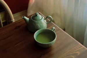 A cozy, close-up photograph captures a light green ceramic tea set resting on the corner of a dark, rustic wooden table. The set includes a small teapot decorated with intricate, embossed floral patterns and a matching wide-rimmed teacup filled to the brim with vibrant, opaque green tea. The scene is illuminated by soft, natural light streaming in from the right, partially filtered by a sheer white curtain in the background, which creates a warm, serene, and inviting atmosphere.