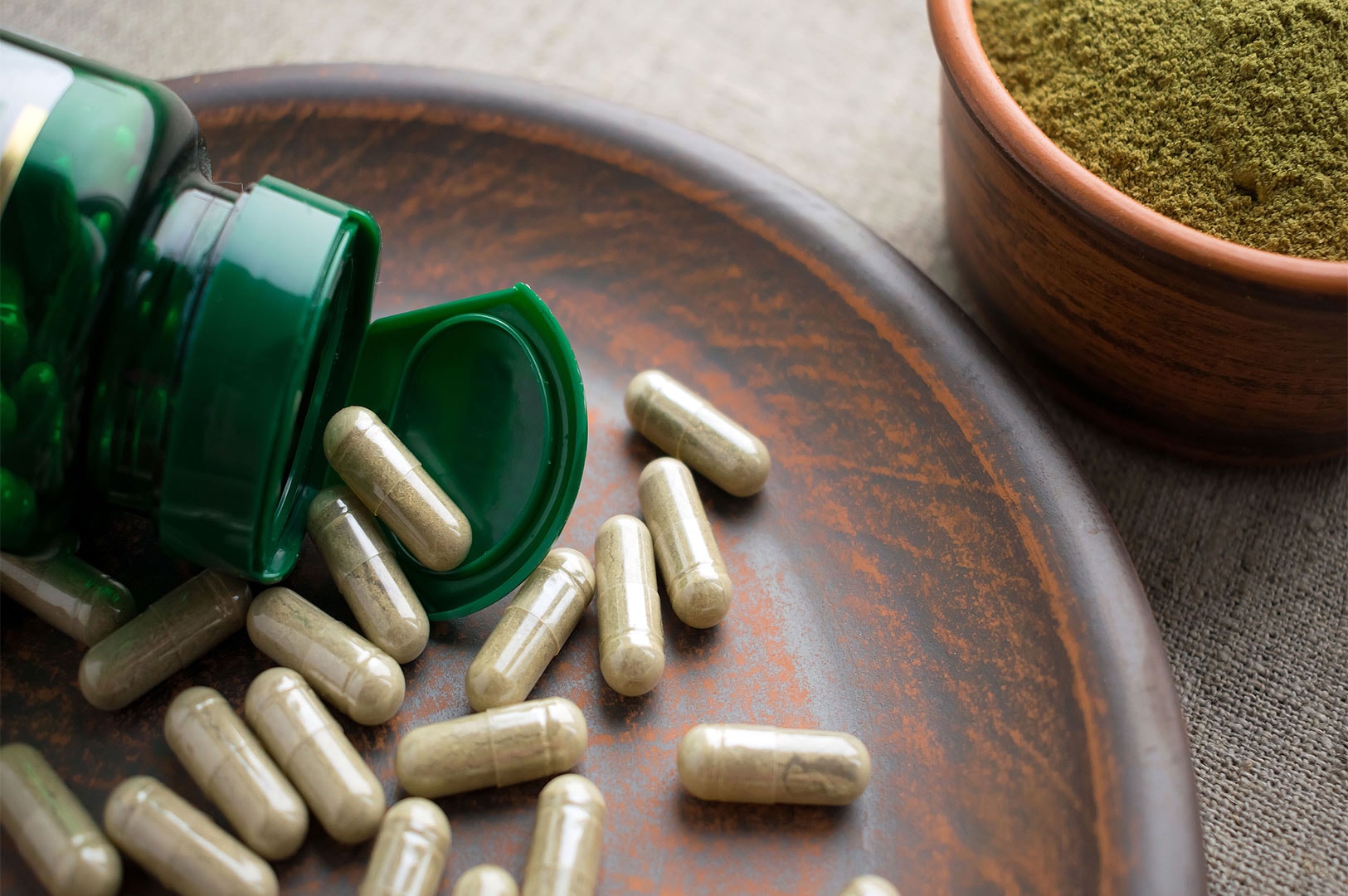 A spilled green plastic bottle with herbal supplement capsules scattered on a brown ceramic plate, with a bowl of green powder (likely matcha or an herbal extract) in the background.