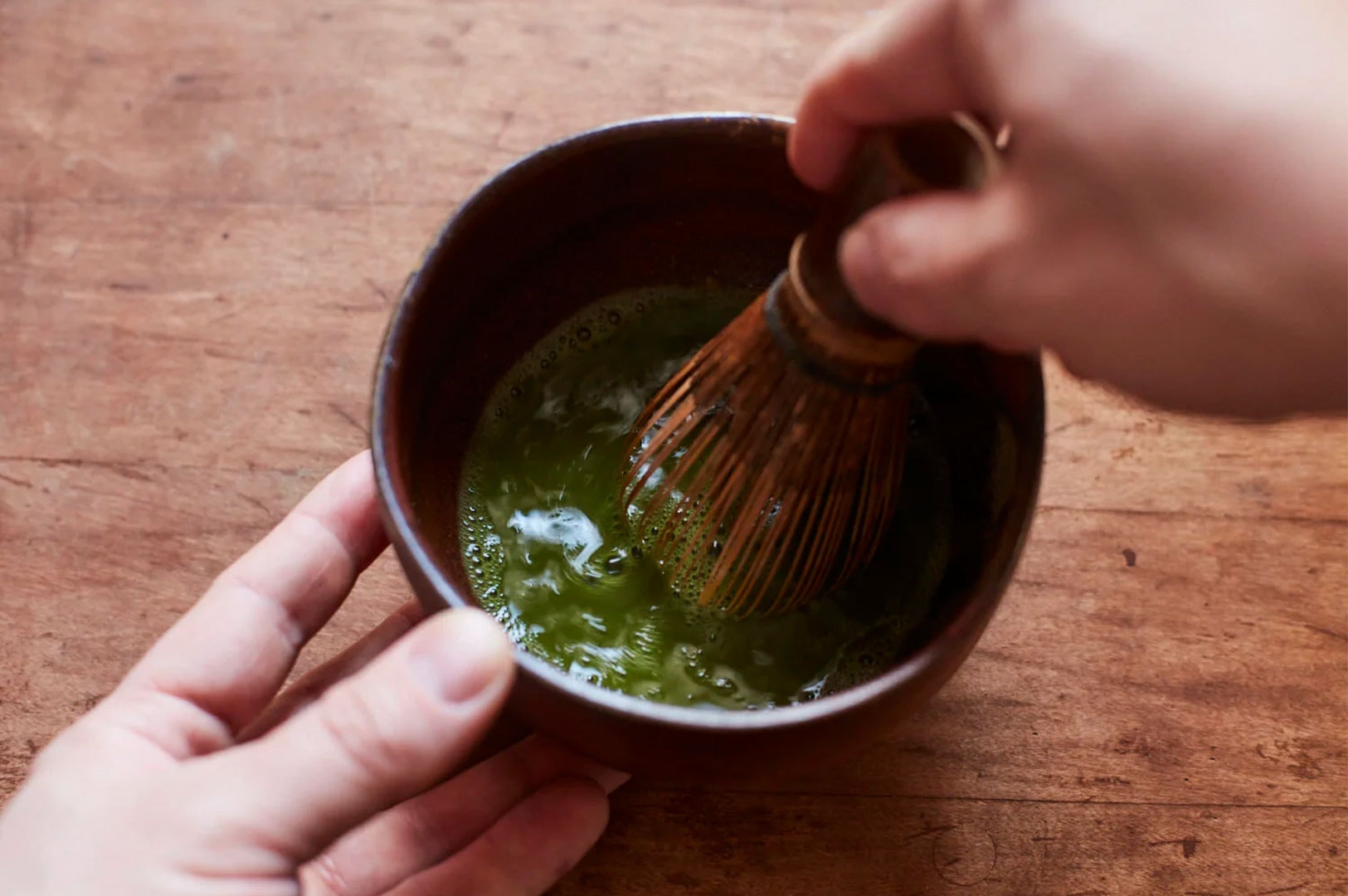 A person's hands whisking bright green matcha tea with a bamboo whisk (chasen) inside a dark wooden bowl.