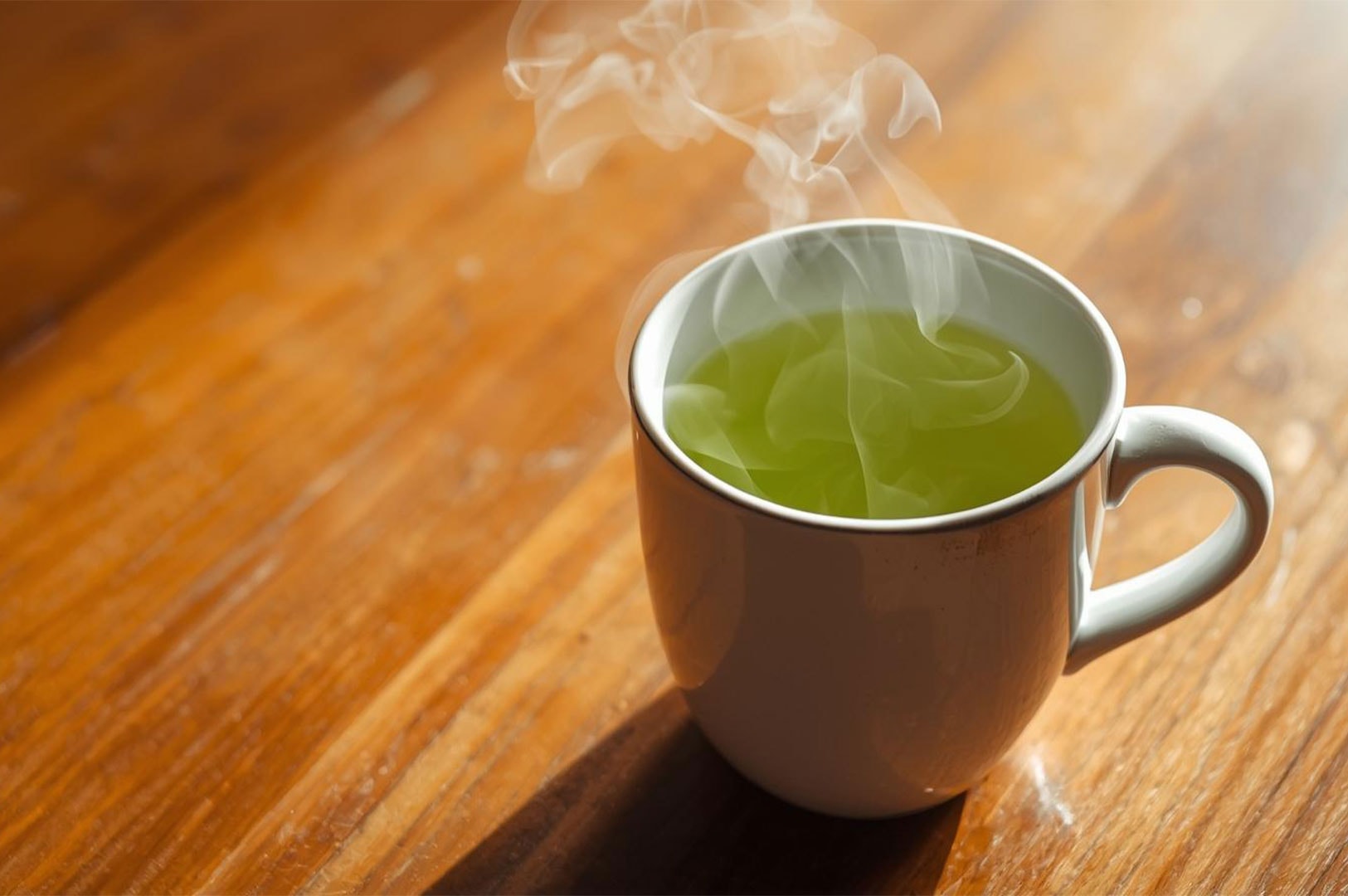 A white ceramic mug filled with bright green, steaming matcha or green tea, sitting on a warm wooden table in natural sunlight.