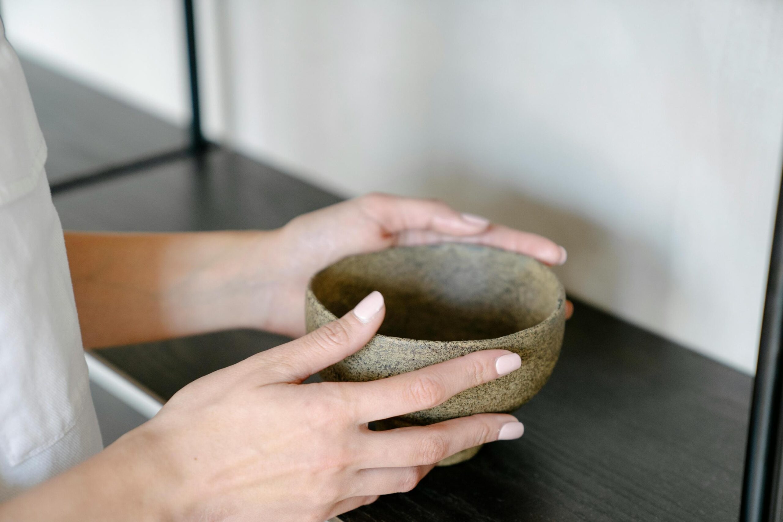 In this minimalist composition, a person with light-colored manicured nails gently holds a textured, earth-toned ceramic bowl with both hands. The bowl is set against a dark shelf and a plain white background, emphasizing a clean and calm aesthetic.