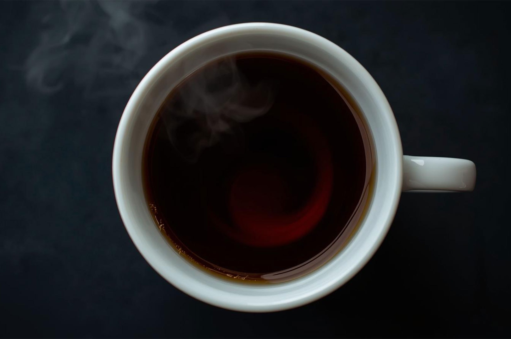 Overhead shot of a dark-brewed black tea in a simple white ceramic mug, with a small curl of steam rising from the liquid.
