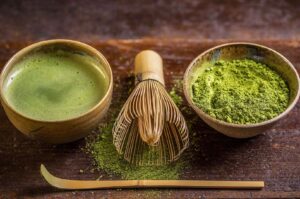 Traditional matcha tea set showing a bowl of prepared matcha, a chasen (whisk), a bowl of vibrant green matcha powder, and a chashaku (bamboo scoop) on a wooden surface.
