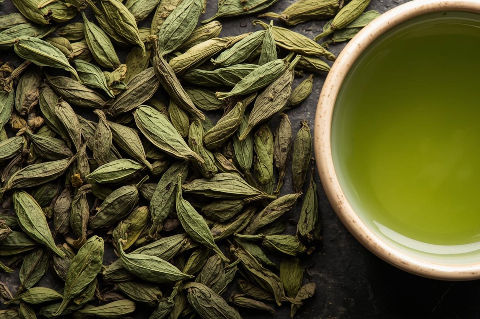 Overhead close-up shot of a cluster of dried green seed pods or husks next to a small bowl filled with bright green tea, possibly matcha.