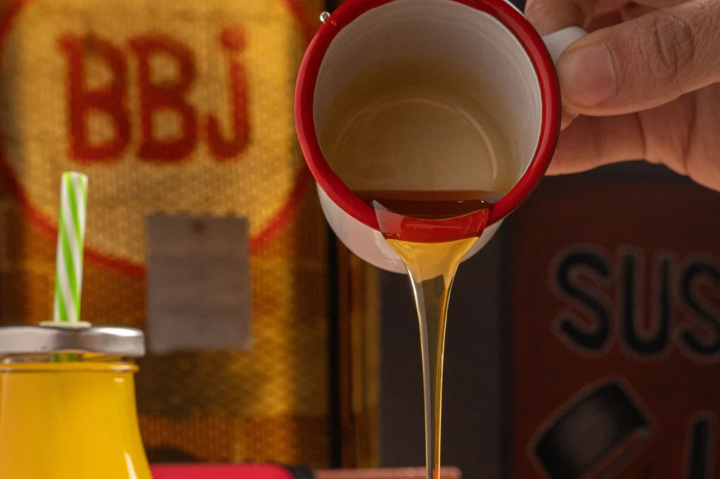 A close-up shot capturing a thick, golden syrup or honey pouring in a smooth stream from a small white enamel mug with a vibrant red rim. A person's hand holds the mug from the right side of the frame, tilting it to drizzle the translucent liquid downward. In the blurry background, a yellow glass bottle with a green and white striped straw sits on the left, positioned in front of a rustic, textured yellow backdrop featuring the red letters "BBJ". To the right, a darker red container with the partial word "SUS" is faintly visible, creating a warm, vintage cafe atmosphere.