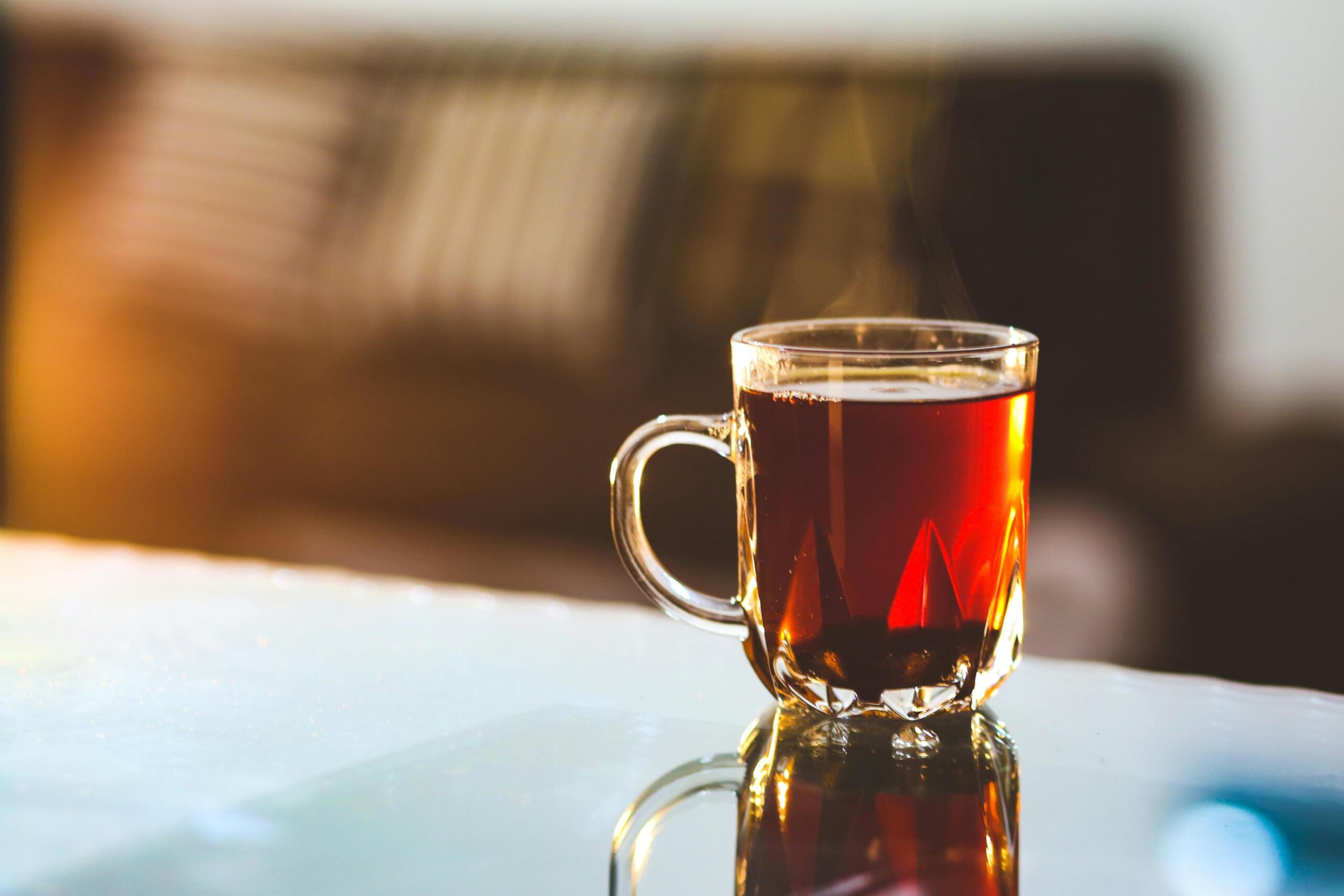 A clear glass mug filled with hot tea rests on a reflective table, illuminated by warm light that highlights the rising steam. Behind the cup, a blurred background features a sofa with striped pillows, creating a cozy and inviting domestic atmosphere.