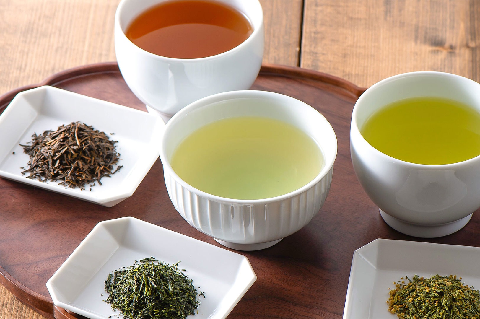 A display of four different tea varieties on a wooden tray, showing three bowls of brewed tea (two green, one dark amber) and three small square dishes of corresponding dry tea leaves (rolled dark, bright green, and genmaicha).