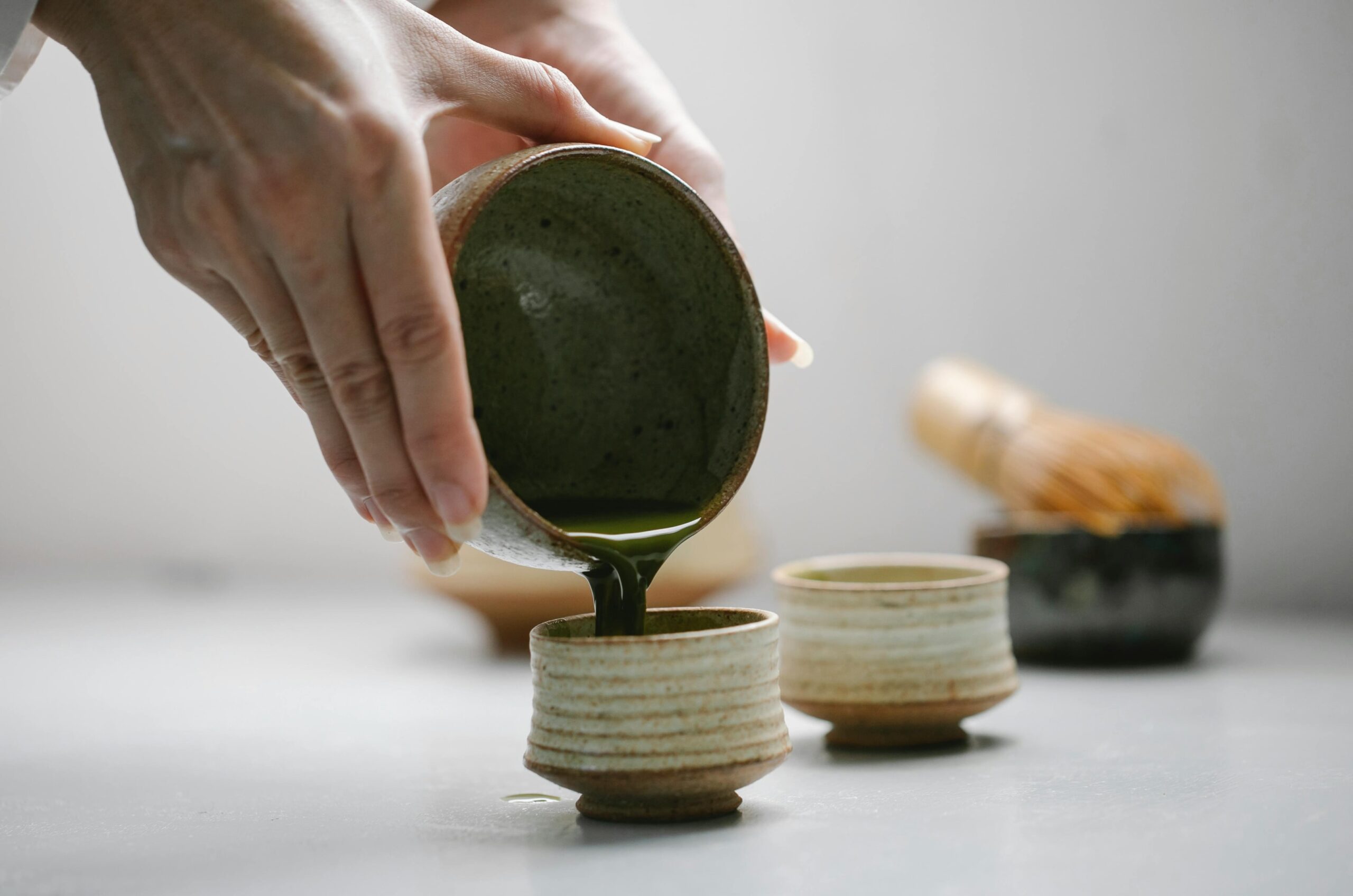  In this minimalist setting, a person carefully pours vibrant green matcha tea from a large ceramic bowl into a smaller, textured teacup. In the soft-focus background, a traditional bamboo whisk sits ready, completing the serene tea preparation scene.
