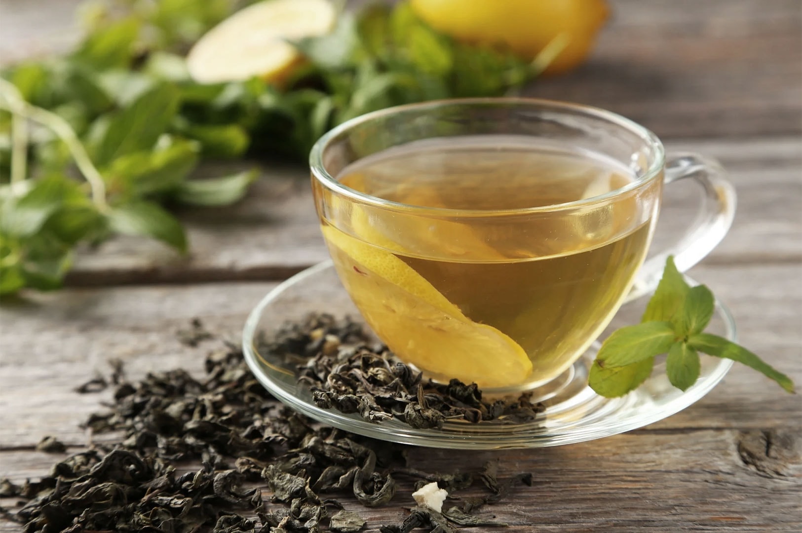 A clear glass teacup with hot tea and a slice of lemon on a saucer, surrounded by scattered loose dried green tea leaves and fresh mint sprigs on a rustic wooden table.