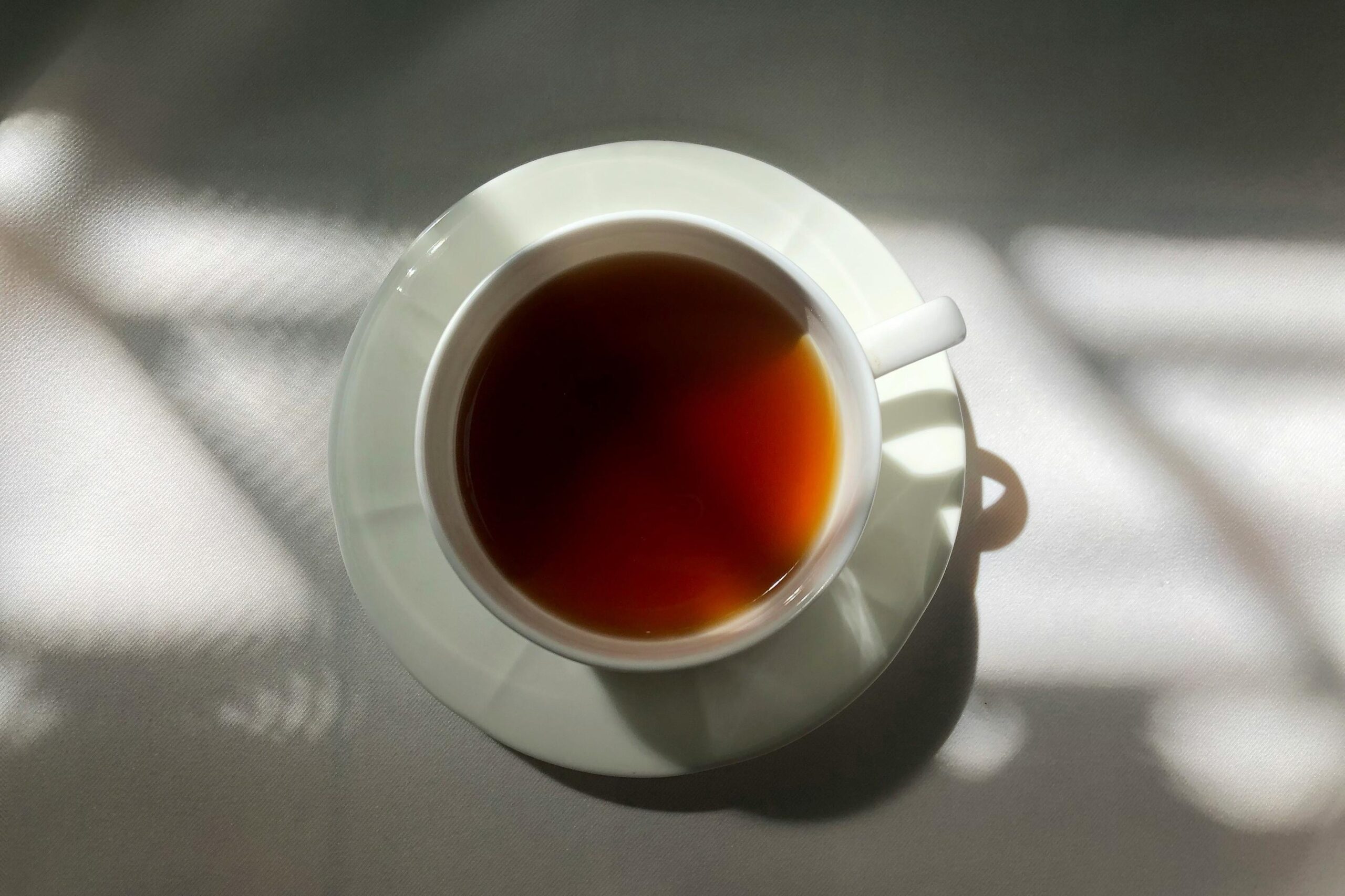 A top-down view captures a classic white teacup filled with amber liquid, resting centrally on a matching saucer. Distinct, patterned shadows stretch across the light surface, creating a high-contrast interplay between the bright highlights and dark silhouettes.