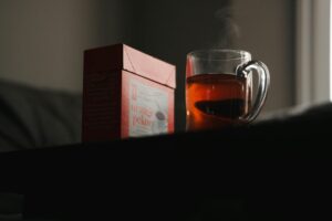 A red box of Orange Pekoe tea sits beside a clear glass mug filled with hot, amber liquid. Steam rises gently from the cup, creating a cozy atmosphere against the moody, dimly lit background.