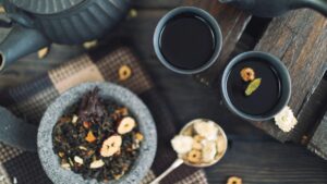 A top-down perspective captures a dark ceramic teapot and two matching cups filled with tea, one of which is delicately garnished with a green cardamom pod and a dried fruit slice. Accompanying the service is a small stone bowl brimming with loose tea leaves and dried botanicals, all arranged on a rustic wooden table with a textured fabric accent.