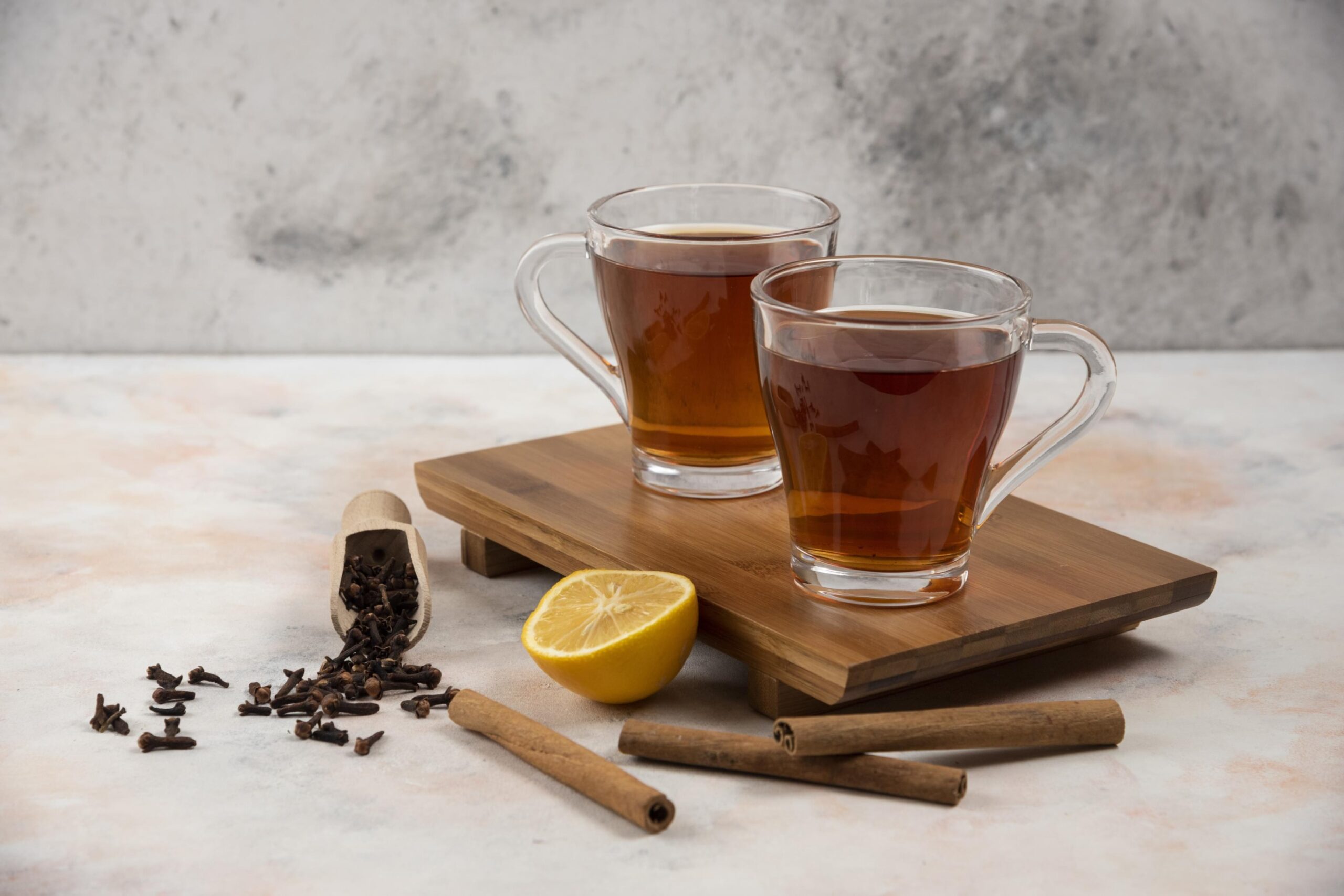Two clear glass mugs filled with tea rest on a small wooden serving board against a textured gray background. The arrangement is garnished with aromatic ingredients, featuring three cinnamon sticks, a fresh lemon half, and a wooden scoop spilling whole cloves onto the surface.