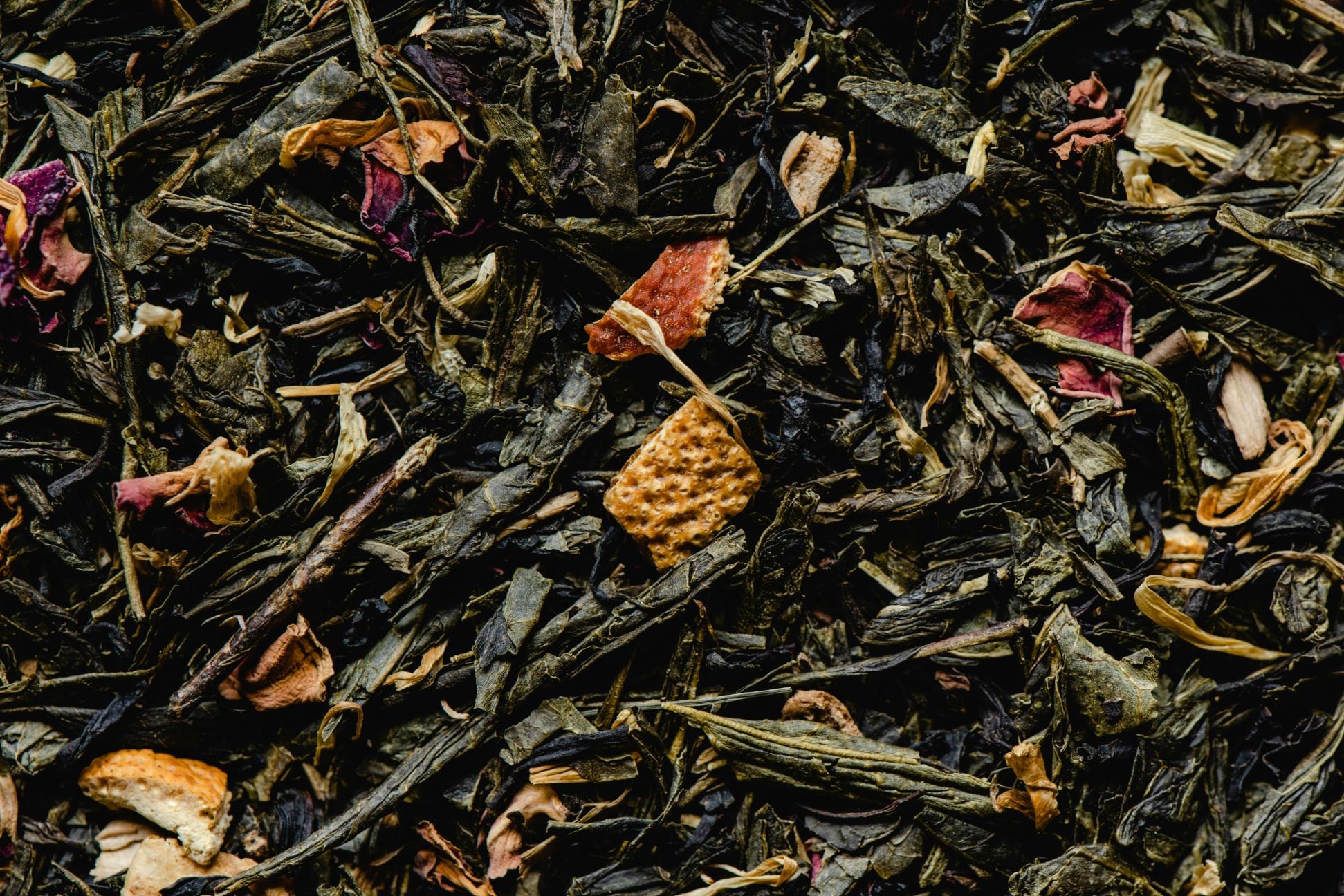 This macro shot showcases a textured blend of loose-leaf green tea mixed with various botanical ingredients. Visible among the dark, twisted leaves are vibrant pink flower petals and small, orange-colored dried citrus peels.