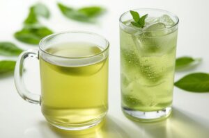 Two glasses of green tea: one glass mug with hot green tea and one tall glass with iced green tea, garnished with mint, on a white surface with out-of-focus green leaves in the background.