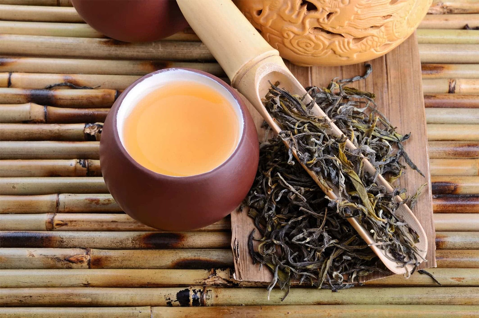 An artistic top-down view of a small brown cup of light amber-colored oolong tea, next to a bamboo scoop overflowing with dry, rolled tea leaves on a mat.